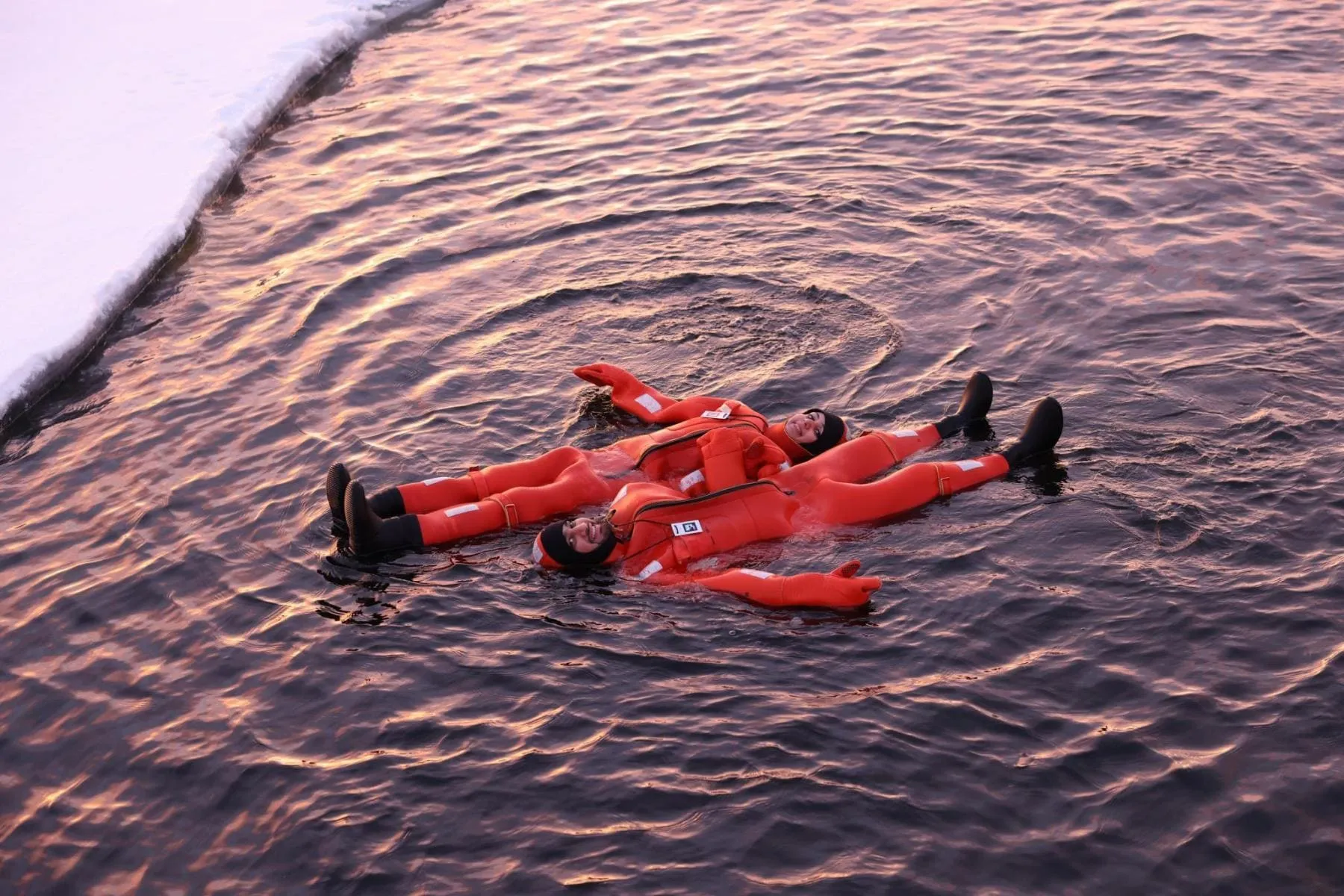Two people wearing orange survival suits floating on their backs in icy water near a snowy shore at sunset.
