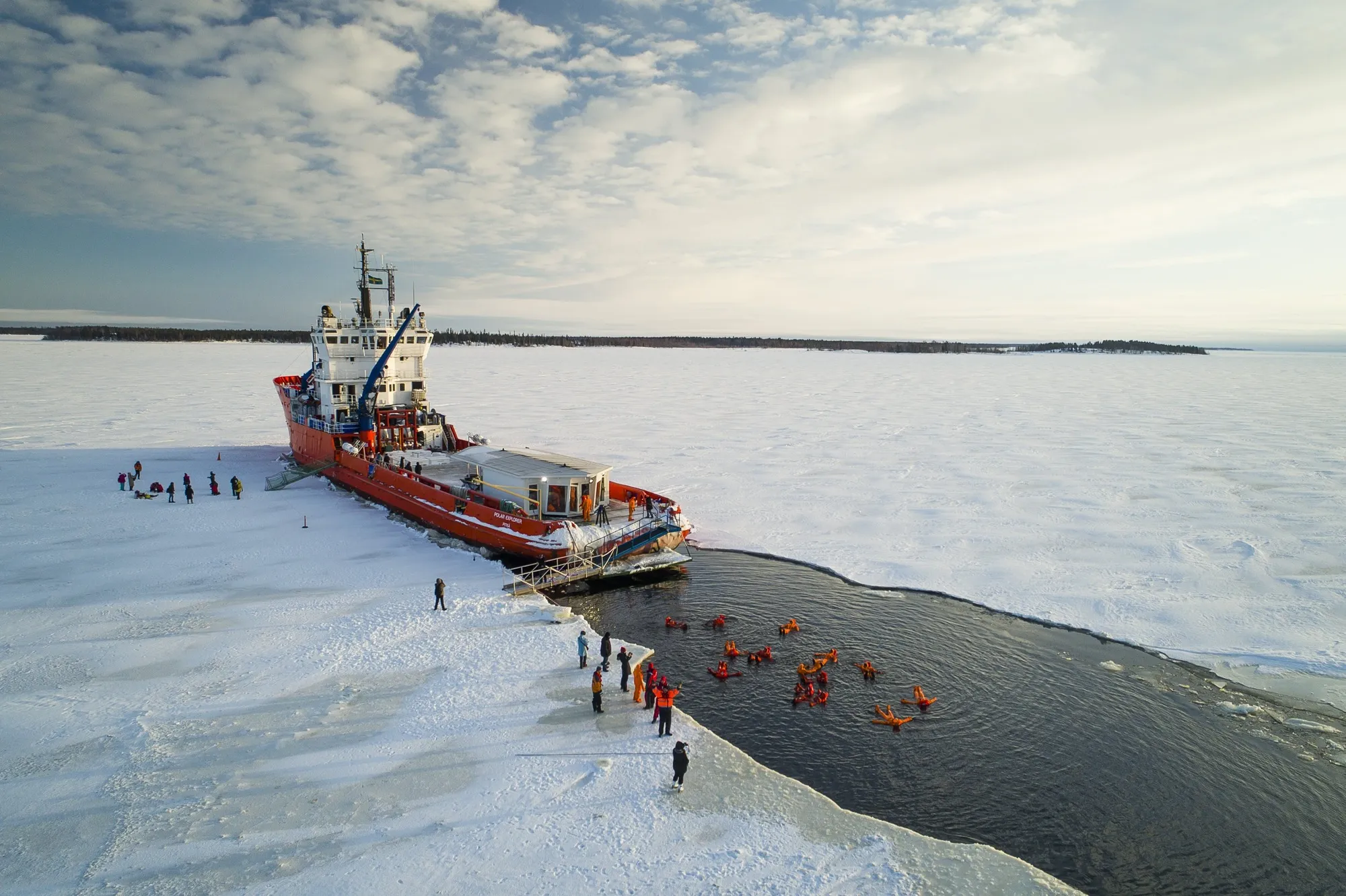 People wearing orange survival suits floating in icy water next to a large red icebreaker ship on a frozen sea with snow-covered ice extending to the horizon.