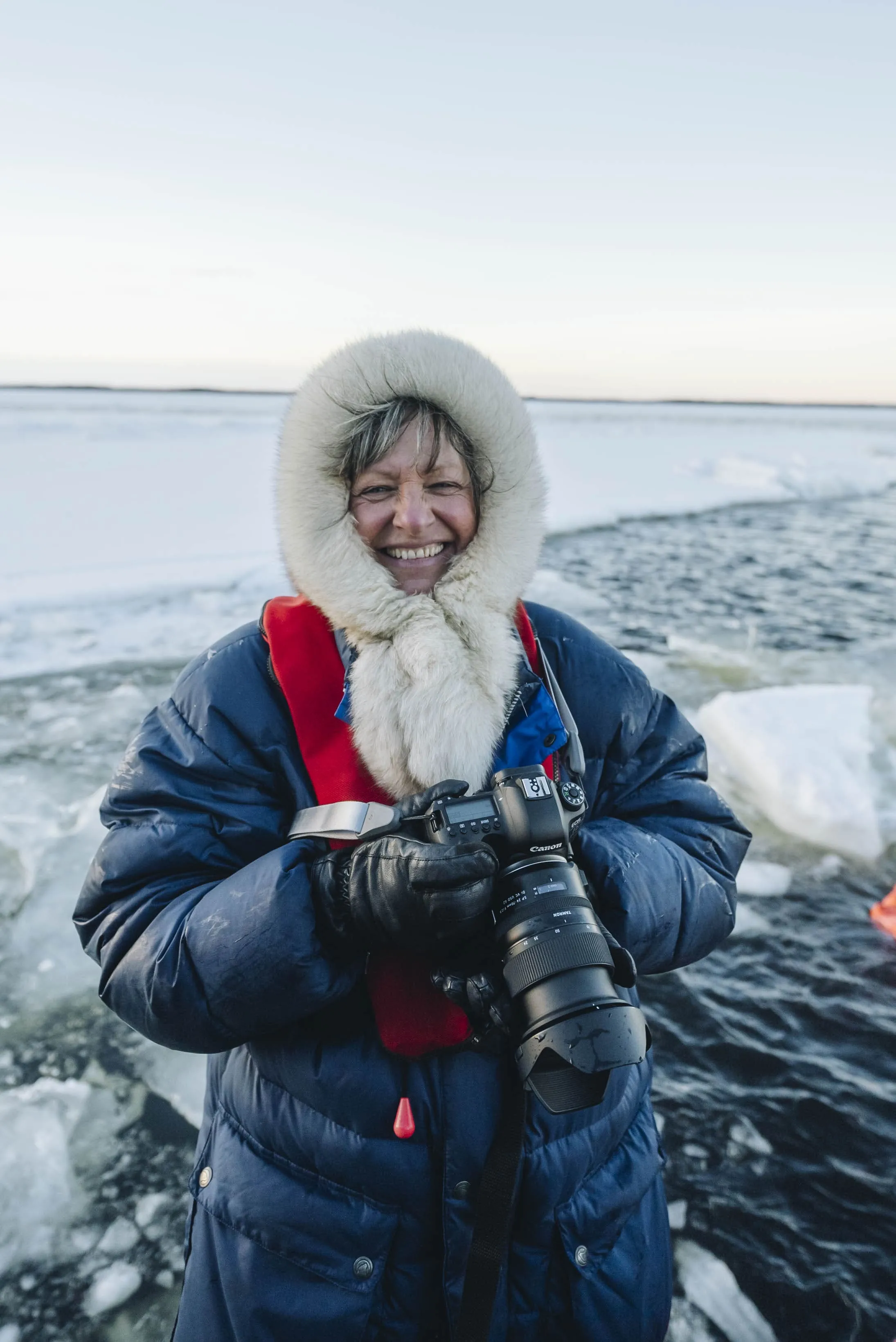 Smiling person in a blue winter coat with fur-lined hood holding a Canon camera near icy water.