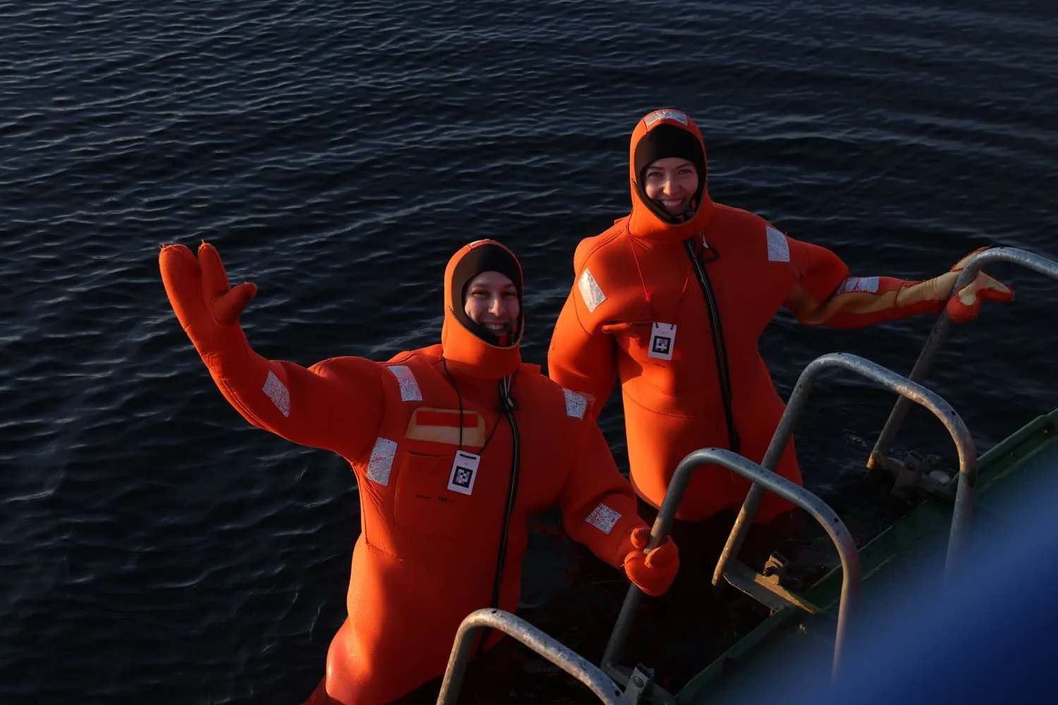 Two people in orange survival suits smiling and holding onto a boat ladder in dark water.