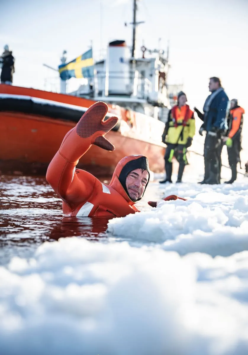 Person in an orange survival suit waving while in icy water near floating ice with a rescue boat and three people in cold-weather gear in the background.
