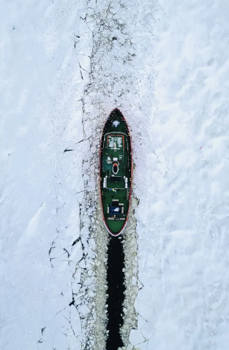 Aerial view of an icebreaker ship cutting through thick sea ice, creating a dark open water path.