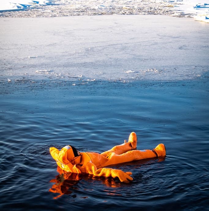 Person wearing a bright orange survival suit floating face-up in dark icy water near a frozen shoreline.