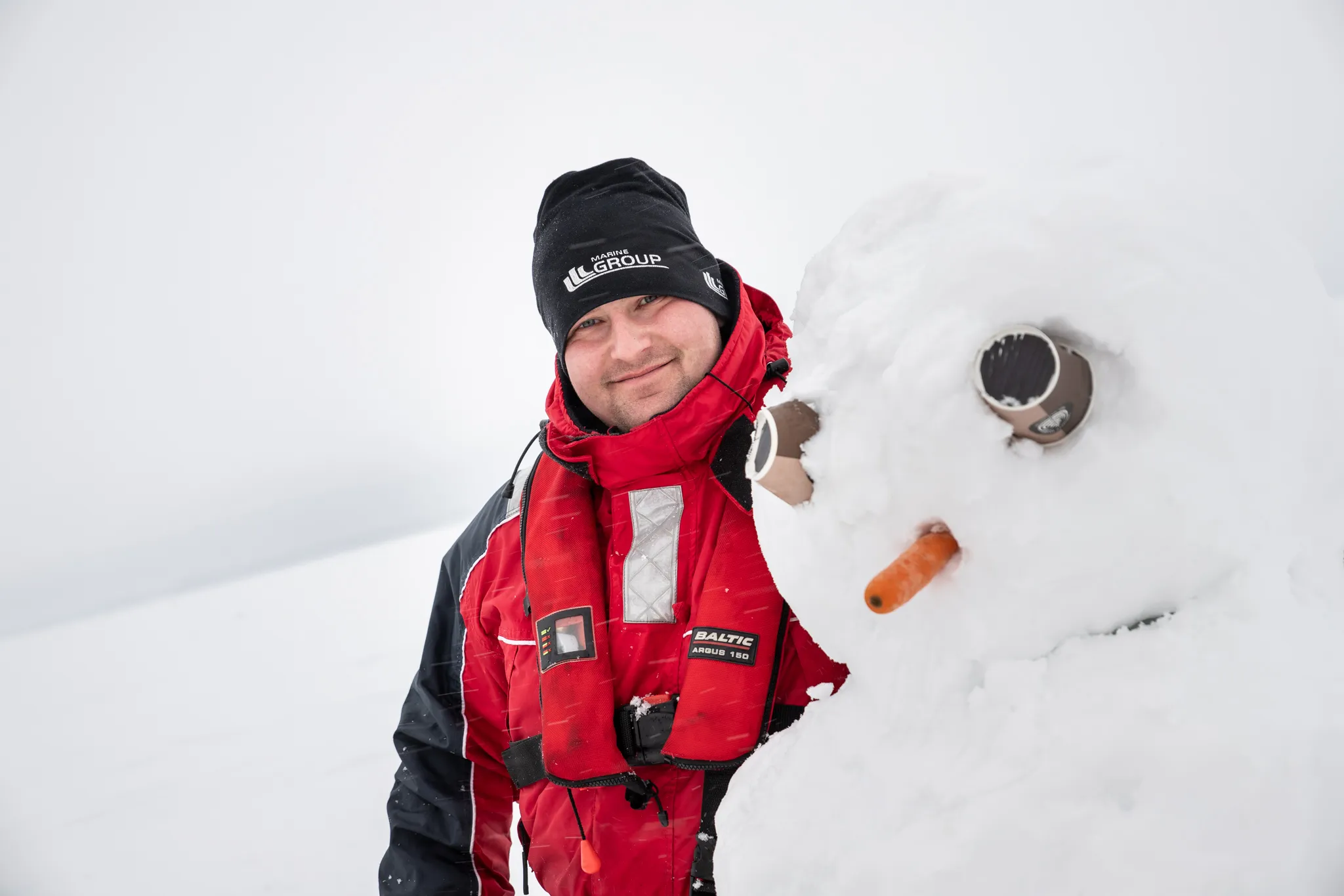 Man in a red and black winter jacket and black hat smiling next to a snowman with a carrot nose and goggles for eyes in a snowy landscape.