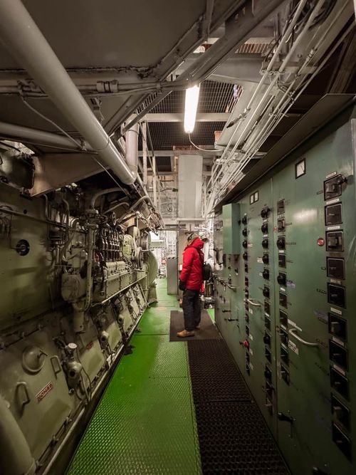 Person in a red jacket and headphones standing inside an industrial engine room with machinery and control panels.