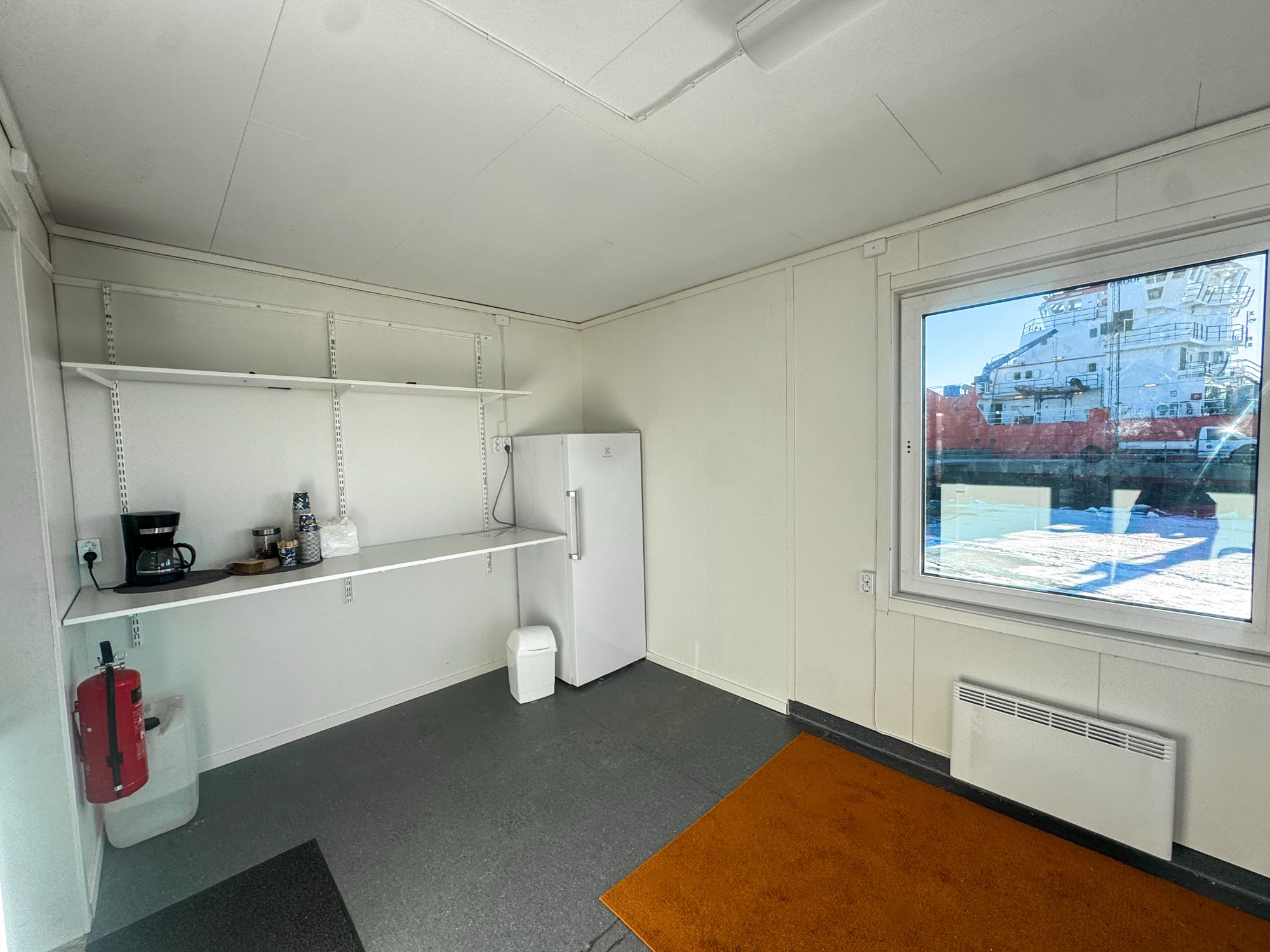 Minimalistic room with a white refrigerator, white shelves holding a coffee maker and cups, a fire extinguisher, a small trash bin, and a window showing an industrial ship outside.
