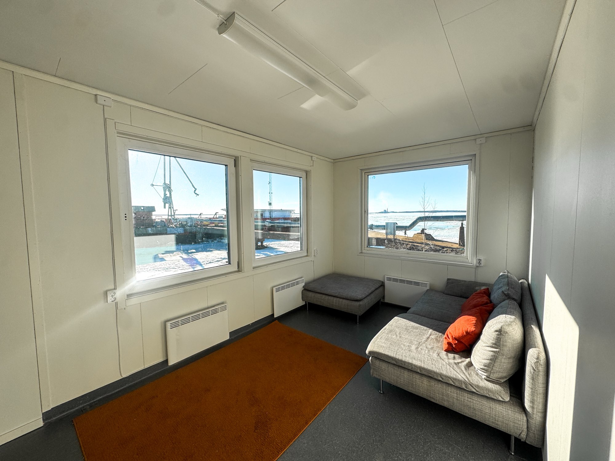 Small sunlit room with three large windows showing an outdoor industrial waterfront, a gray sectional sofa with orange pillows, matching ottoman, and an orange rug on the floor.
