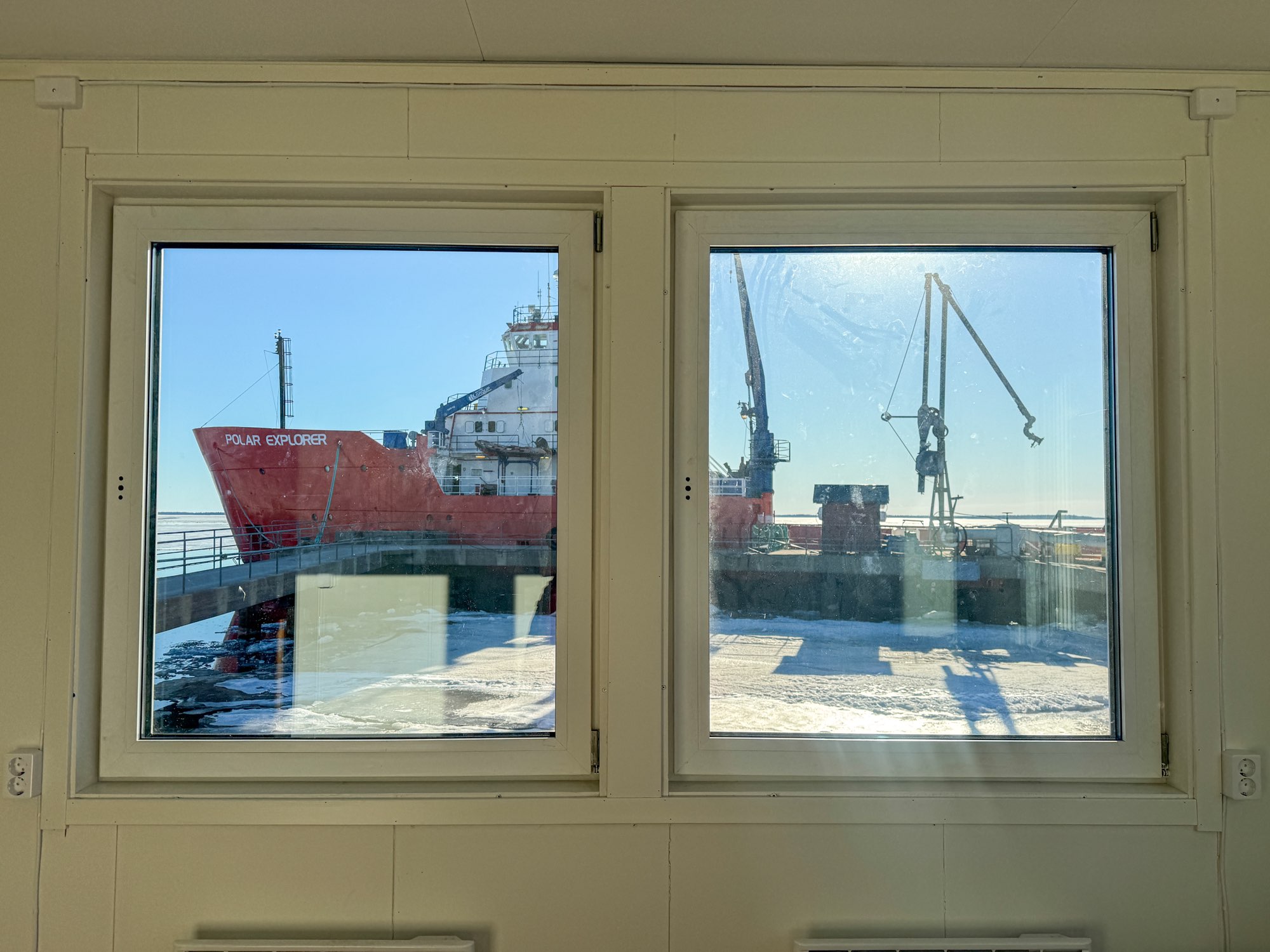 View through two windows showing a large red ship named Polar Explorer docked next to a snowy pier with cranes.