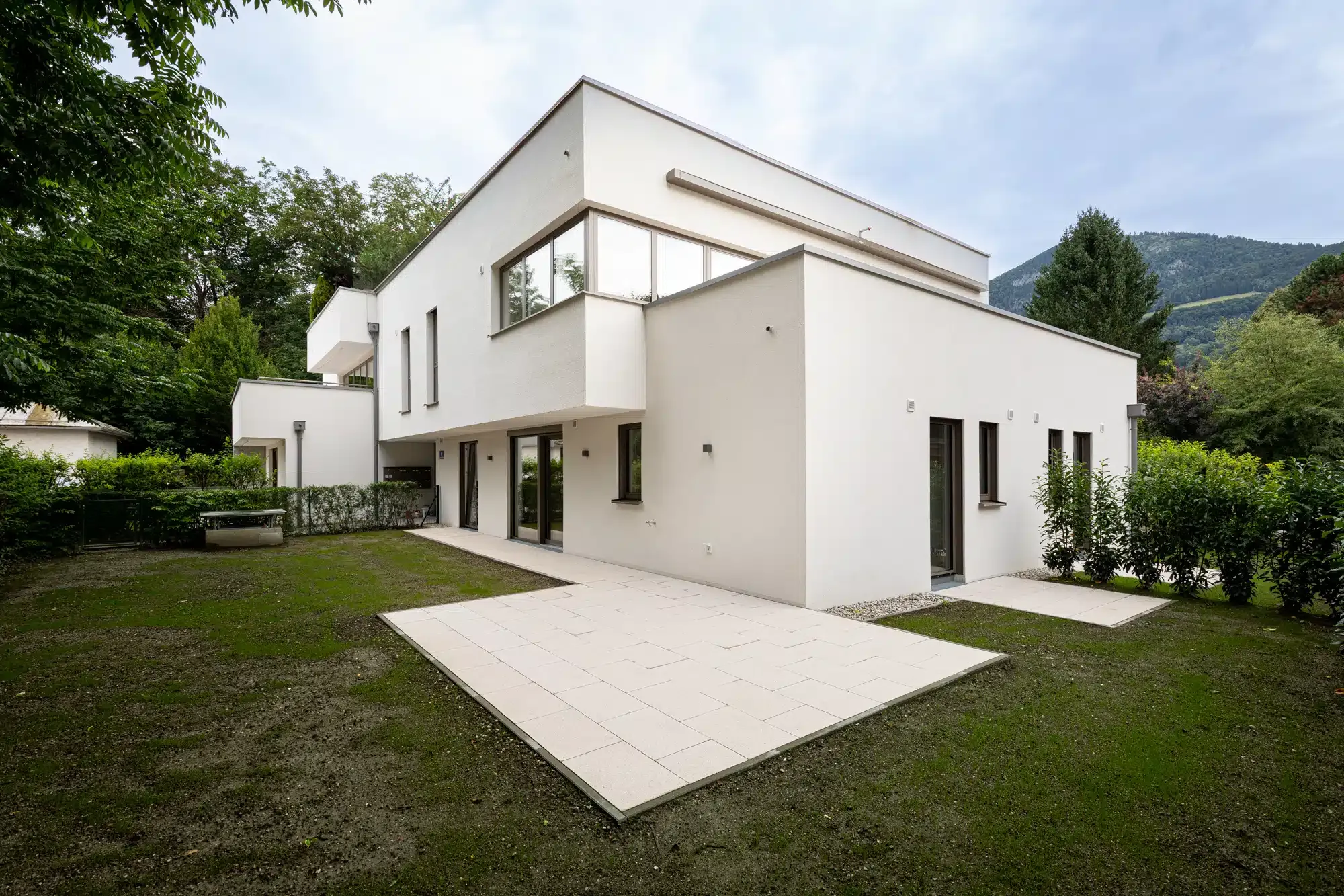 Zweistöckiges Wohngebäude mit weißer Fassade, großen Fenstern und Terrasse in grüner Umgebung