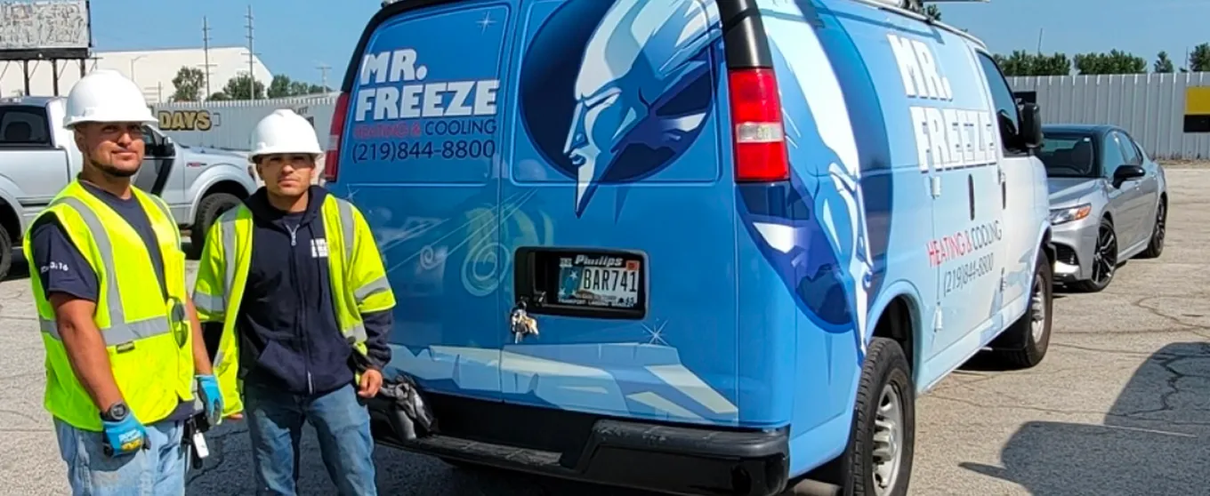 Two men wearing white hard hats and yellow safety vests standing beside a blue Mr. Freeze heating and cooling service van in a parking lot.