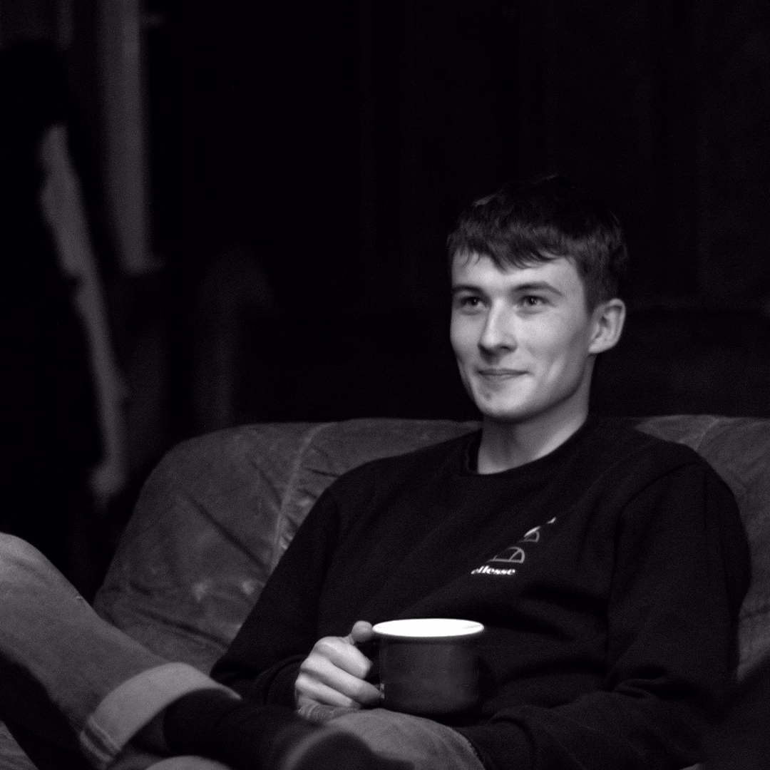 Young man sitting on a couch, holding a mug and smiling slightly in a relaxed pose.