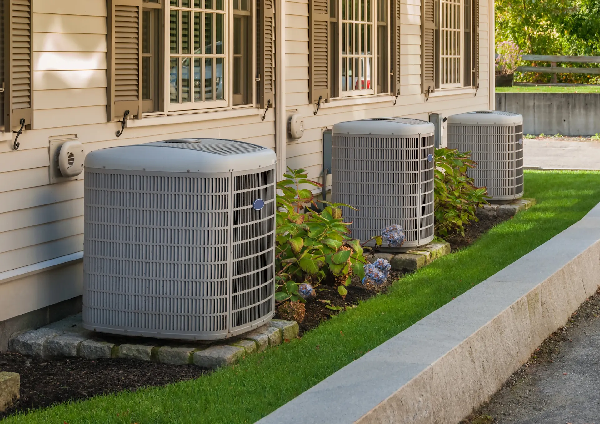 Three outdoor air conditioning units installed on stone slabs along the side of a house with beige siding and shuttered windows.