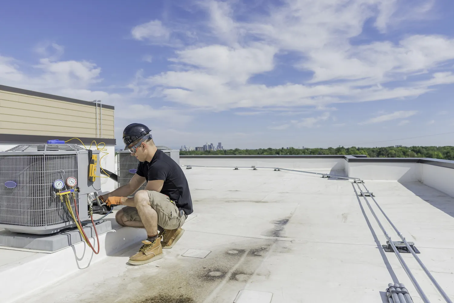 Technician in a black hard hat and gloves repairing rooftop HVAC units on a sunny day.