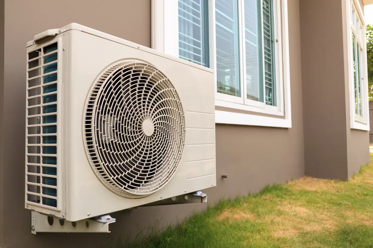 Outdoor air conditioning unit mounted on the exterior wall of a house with beige siding and white-framed windows.