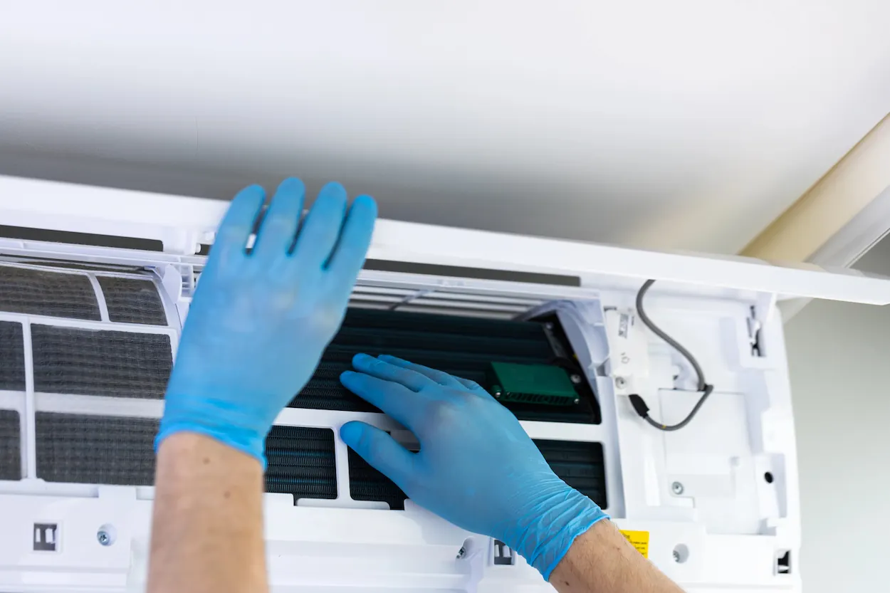 Person wearing blue gloves servicing the interior components of a wall-mounted air conditioning unit.
