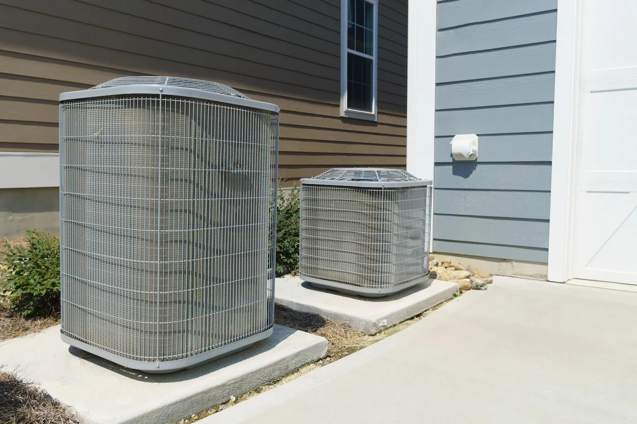 Two outdoor air conditioning units installed on concrete slabs beside a house with brown and blue siding.