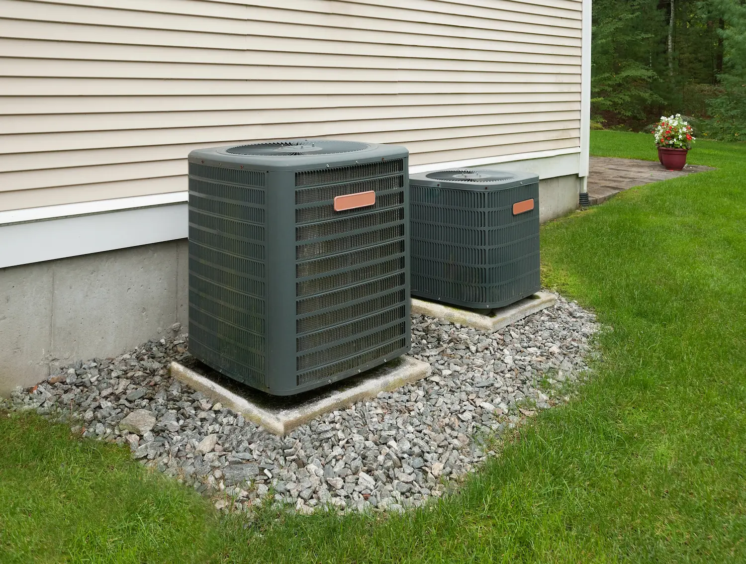 Two outdoor HVAC units beside a beige house on gravel surrounded by green grass.