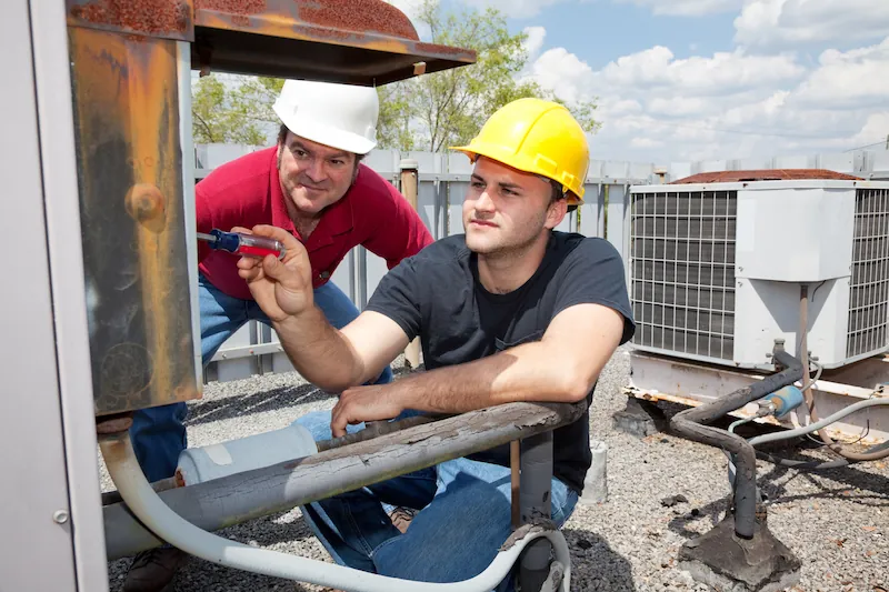 Two workers wearing hard hats inspecting and repairing outdoor HVAC units.