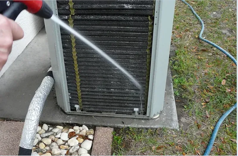 Hand using a pressure washer to clean the coils of an outdoor air conditioning unit.