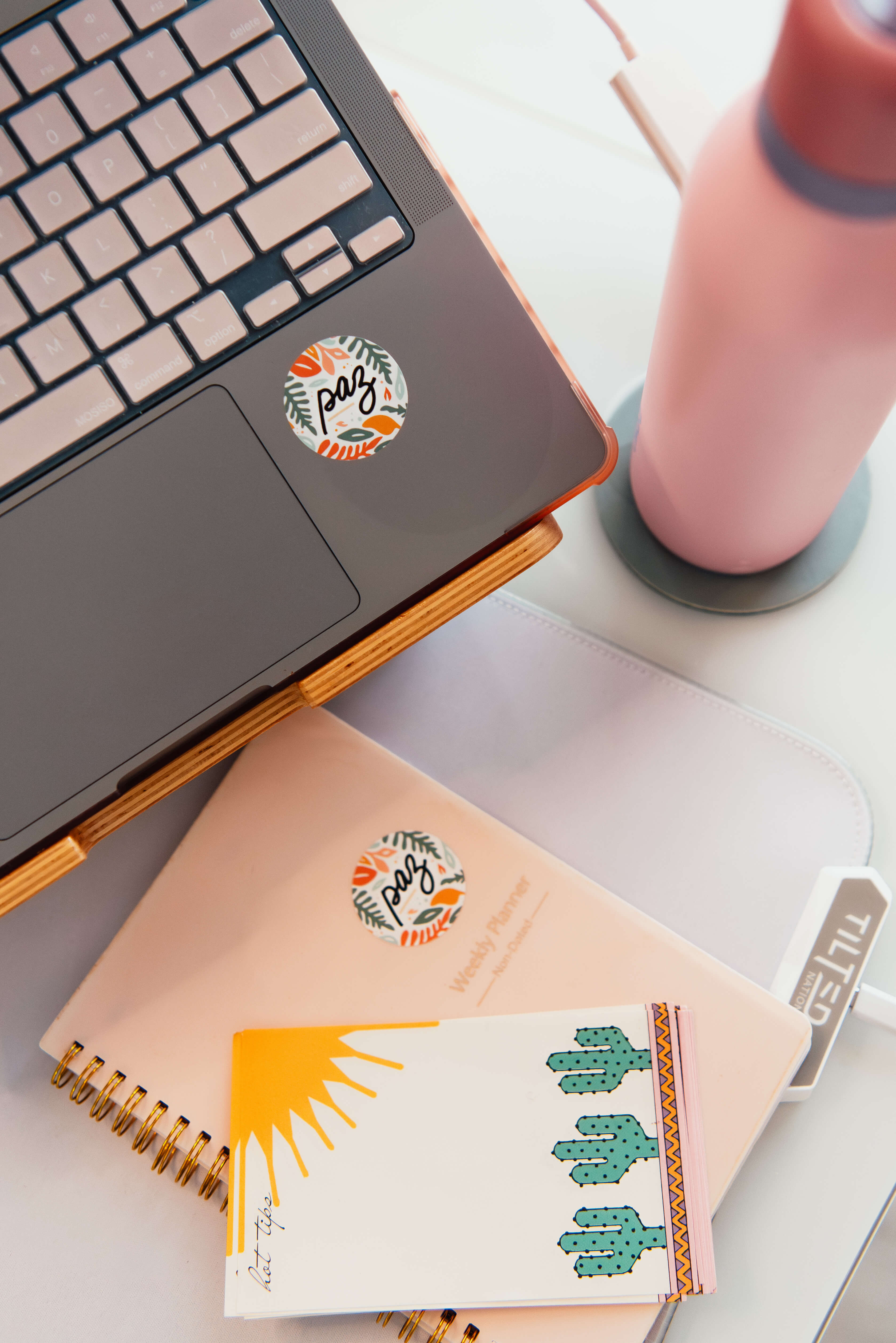 Partial view of a laptop with a round 'paz' sticker next to a pink weekly planner and a notepad featuring a sun and cacti design on a white desk with a pink water bottle.