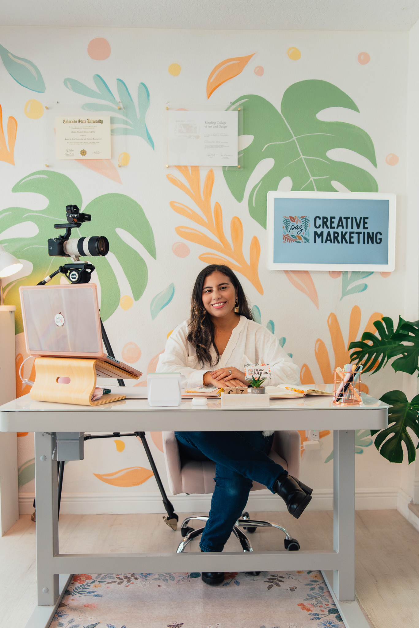Smiling woman sitting at a desk in a colorful office with creative marketing sign and camera on tripod.