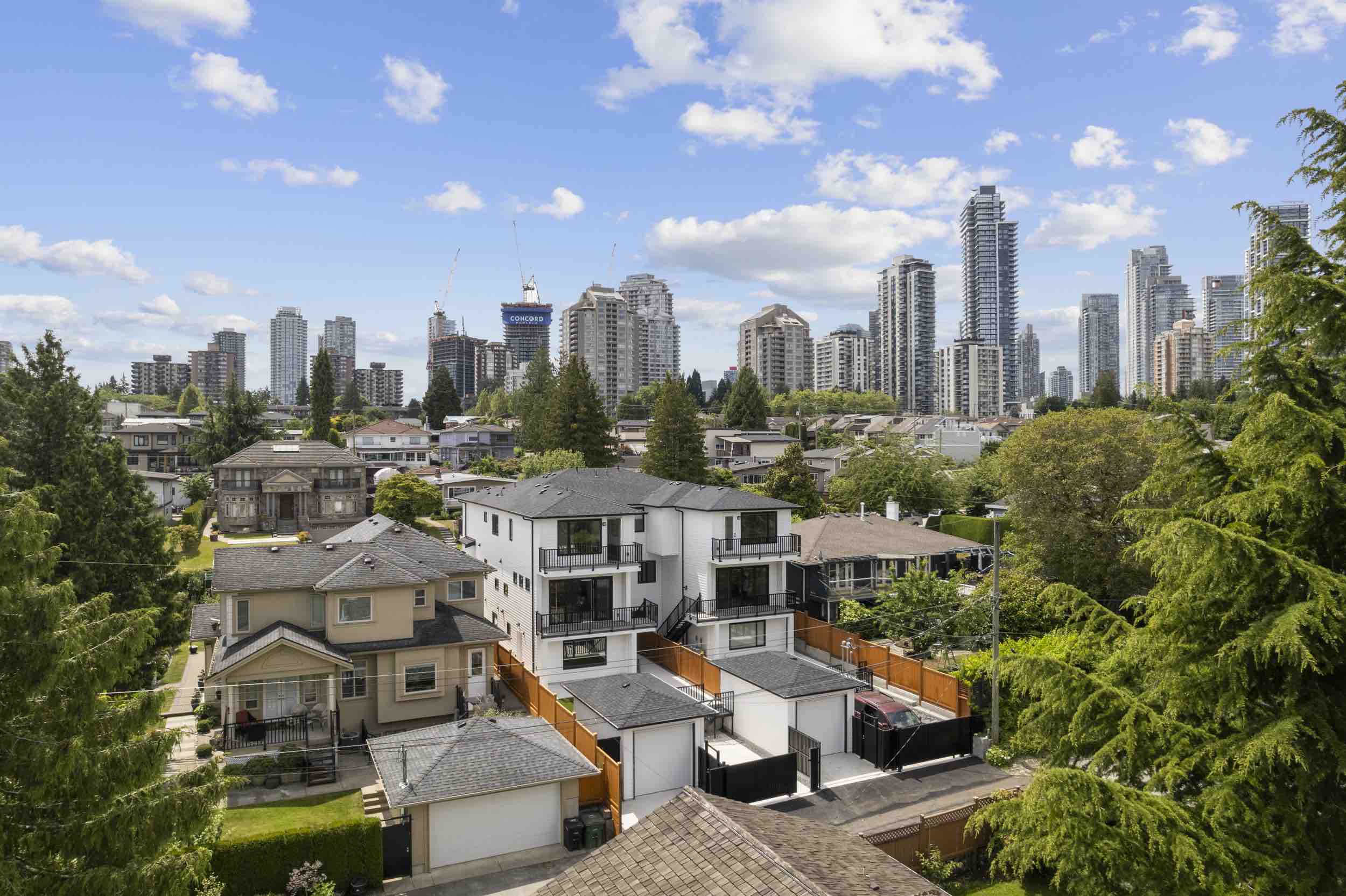 Residential neighborhood with modern multi-story houses in the foreground and a city skyline with high-rise buildings under a blue sky with scattered clouds.