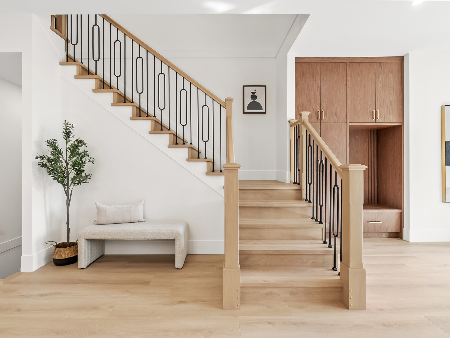 Built by Asthan Homes in Burnaby, BC, Wooden staircase with black metal balusters, a white bench with a cushion, a potted plant, and built-in wooden storage cabinets in a bright modern interior.