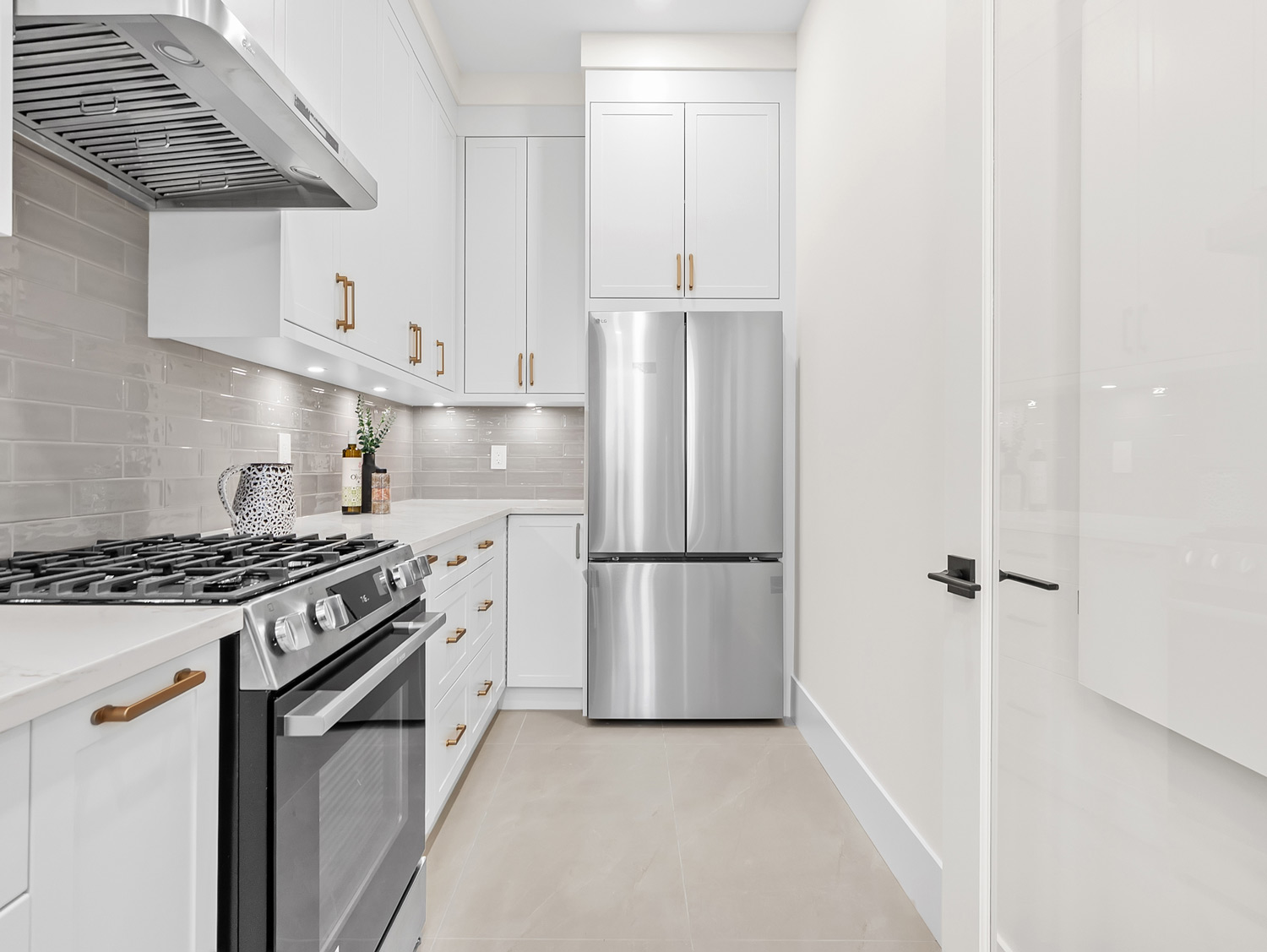 Built by Asthan Homes in Burnaby, BC, kitchen with stainless steel refrigerator, gas stove, white cabinets with gold handles, and beige tiled floor.
