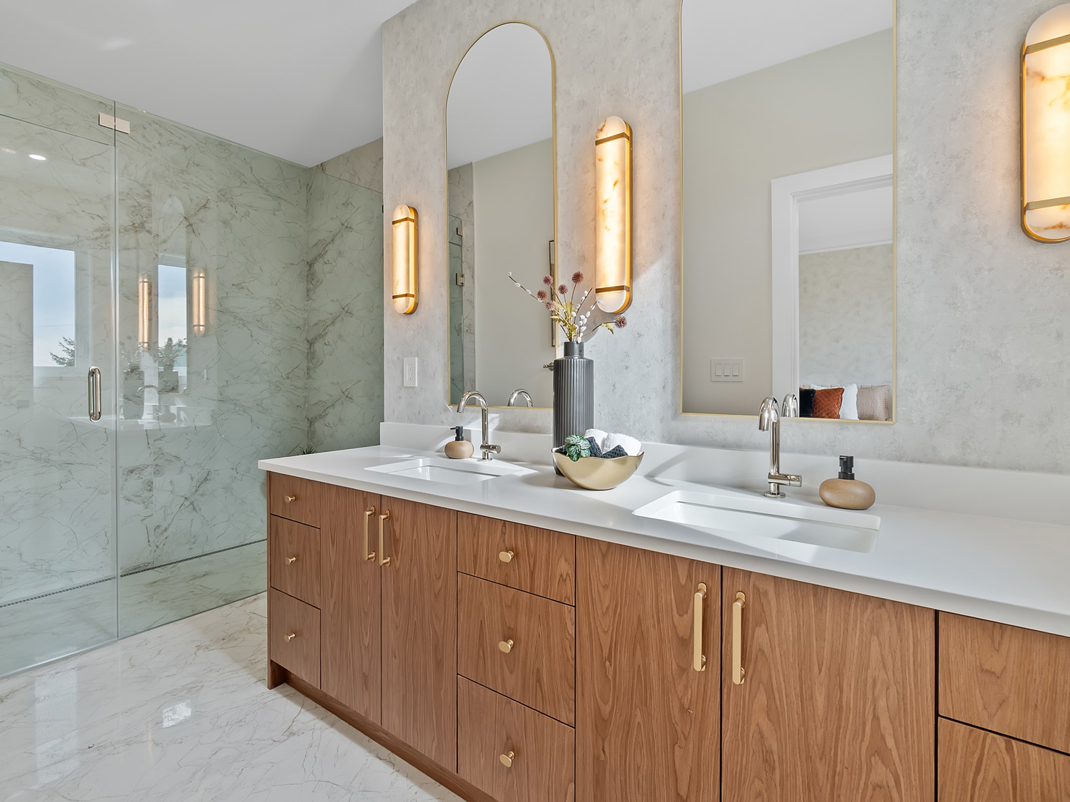 Built by Asthan Homes in Burnaby, BC, bathroom with dual wooden vanity, white countertop, two rectangular sinks, arched and rectangular mirrors, and glass-enclosed marble shower.