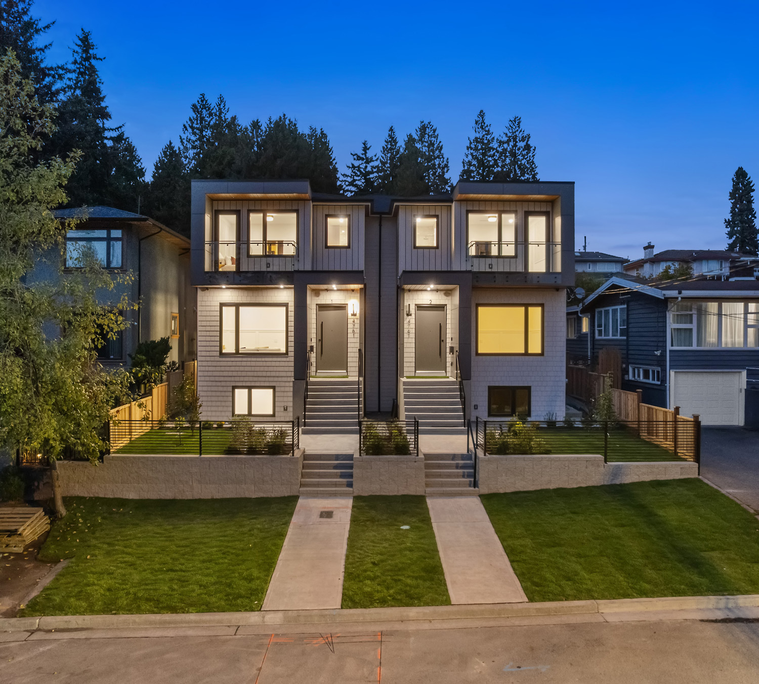 Built by Asthan Homes in Burnaby, BC, duplex house at dusk with illuminated windows, two front entrances, and neatly manicured lawns.