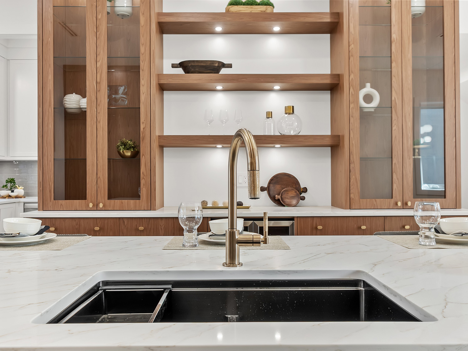 Built by Asthan Homes in Burnaby, BC, kitchen sink with brass faucet set in a white marble countertop, with wooden cabinets and shelves in the background.
