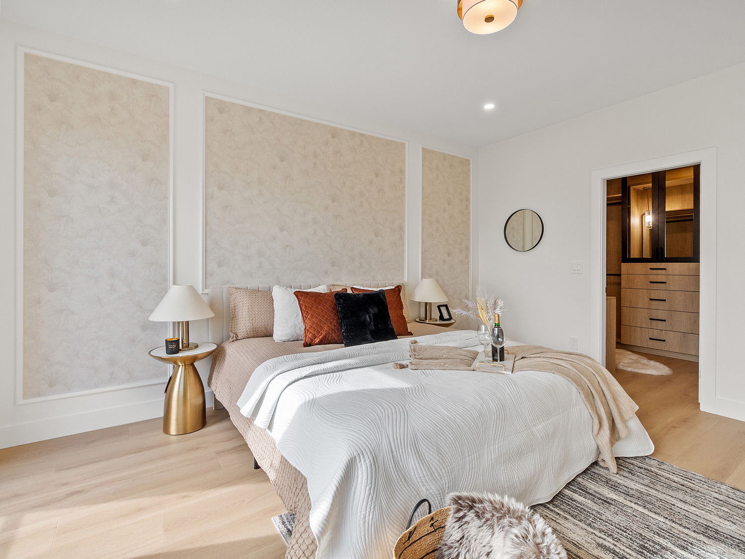Built by Asthan Homes in Burnaby, BC, bedroom with a bed featuring white and beige bedding, decorative pillows, gold nightstands with lamps, and a doorway leading to a walk-in closet.