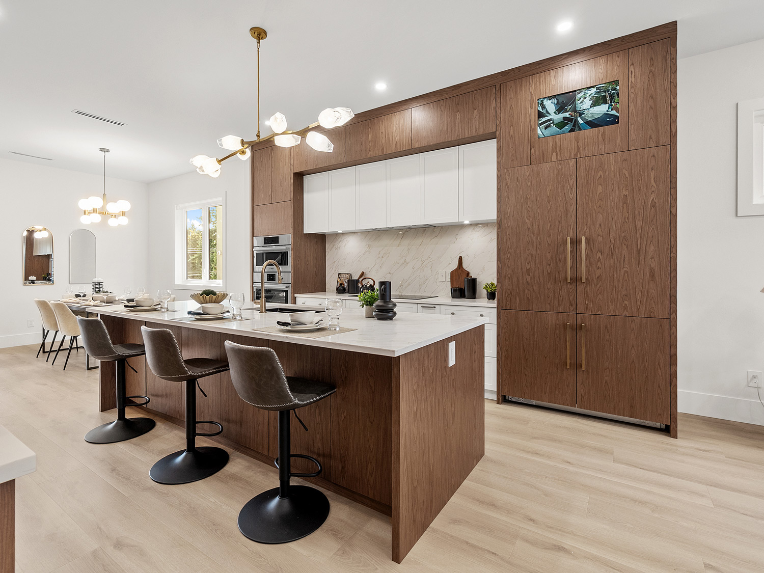 Built by Asthan Homes in Burnaby, BC, modern kitchen with a large wooden island, white countertop, three black bar stools, wood cabinetry, built-in appliances, and two pendant lights.