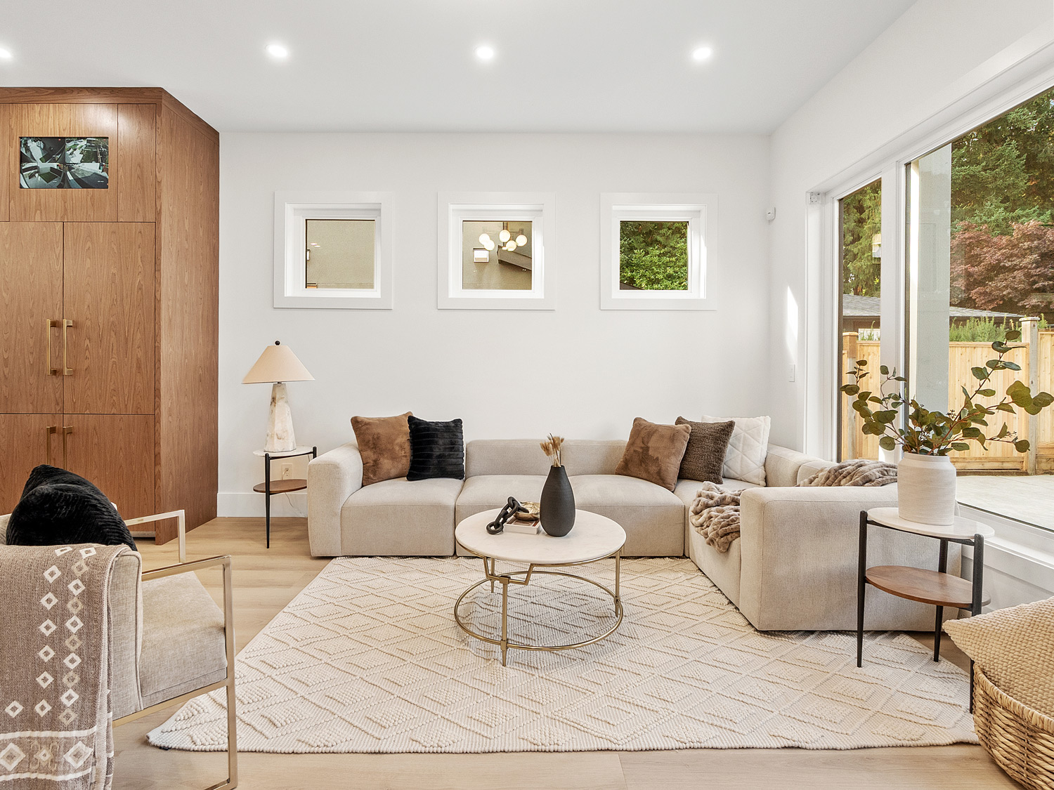 Built by Asthan Homes in Burnaby, BC, living room with beige sectional sofa, gold-trimmed round coffee table, side tables with lamps and vase, and large windows overlooking a fenced backyard.