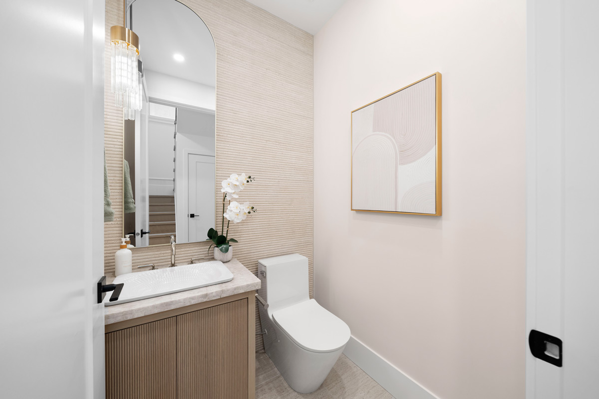 Built by Asthan Homes in Burnaby, BC, bathroom with a beige vanity, white rectangular sink, tall arched mirror, white toilet, and abstract artwork on a light wall.