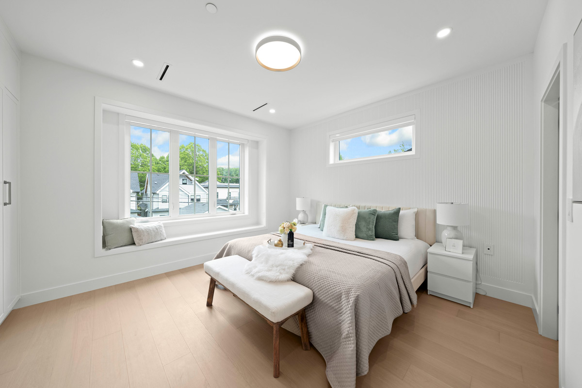 Built by Asthan Homes in Burnaby, BC, bright bedroom with large window seat, king-size bed with beige quilt and green and white pillows, white bench, and nightstands with lamps.