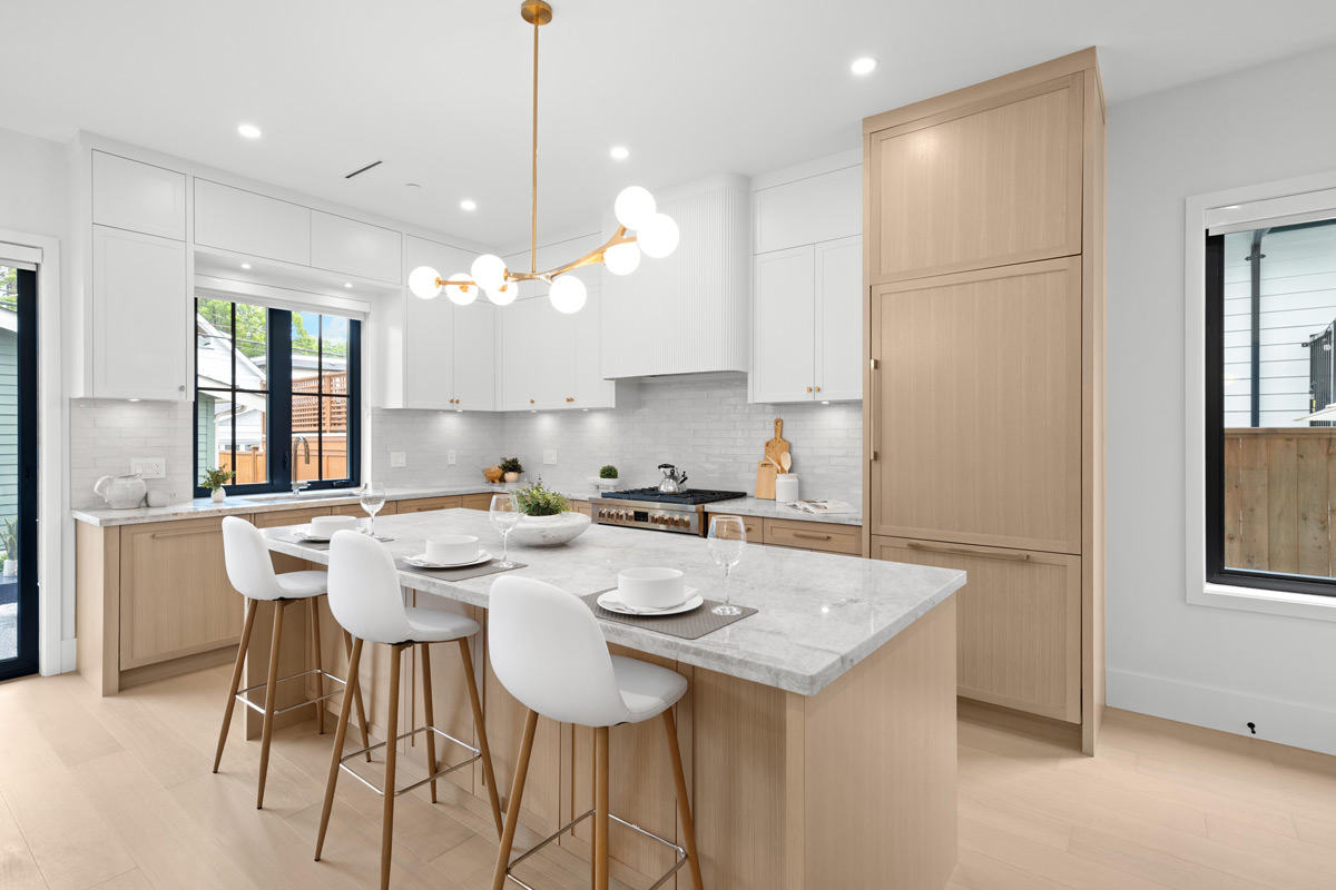 Built by Asthan Homes in Burnaby, BC, kitchen with a large marble island, four white bar stools with wooden legs, white upper cabinets, light wood lower cabinets, and a brass chandelier with globe lights.