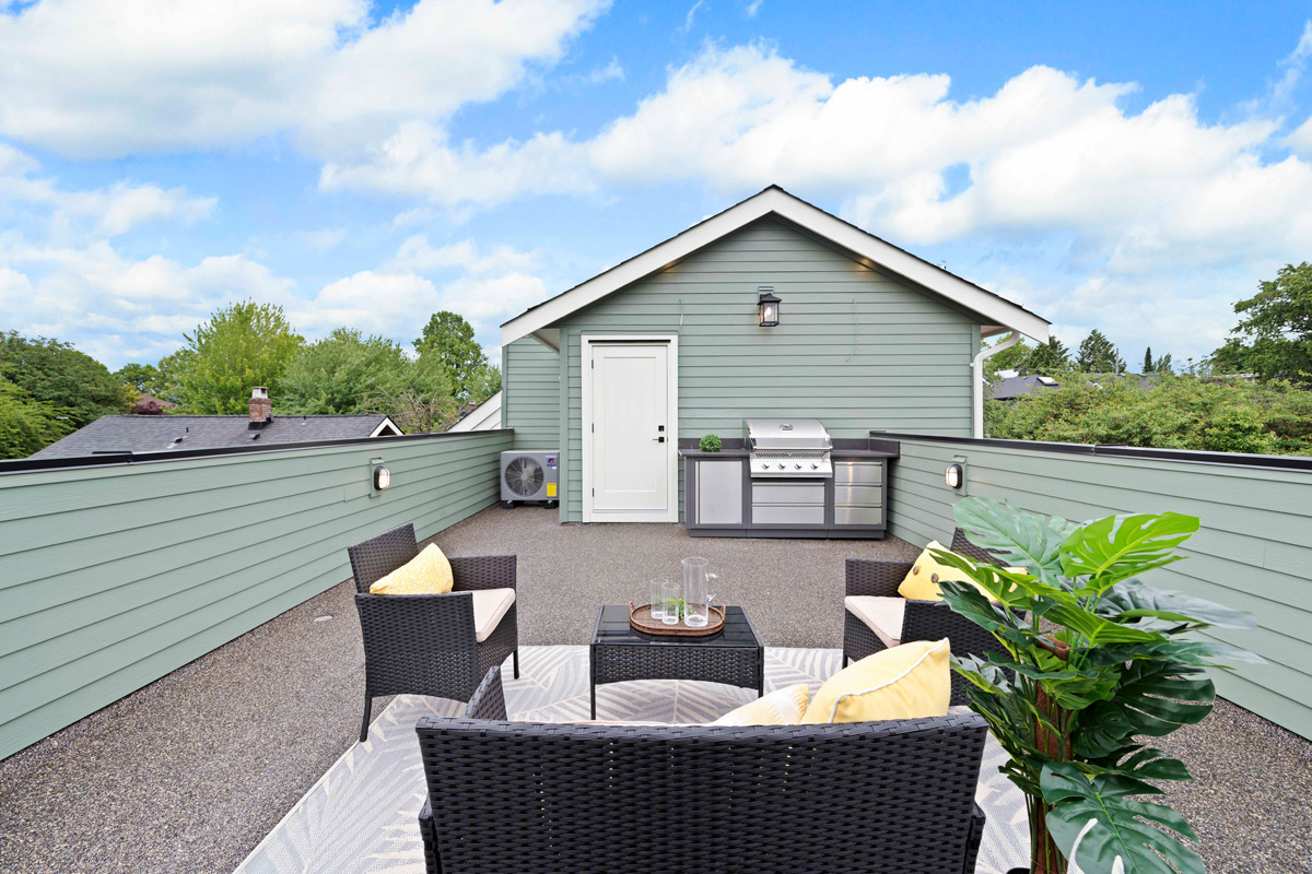 Built by Asthan Homes in Burnaby, BC, outdoor patio with black wicker furniture including sofa and chairs with yellow cushions, a coffee table with glass pitchers, a potted plant, and a built-in stainless steel grill against a green house wall under a blue sky with clouds.