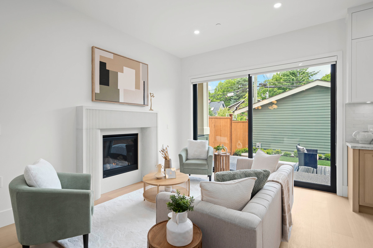 Built by Asthan Homes in Burnaby, BC, bright living room with beige sofa, green armchairs, modern fireplace, round wooden coffee table, and large sliding glass doors overlooking a backyard with green fence.