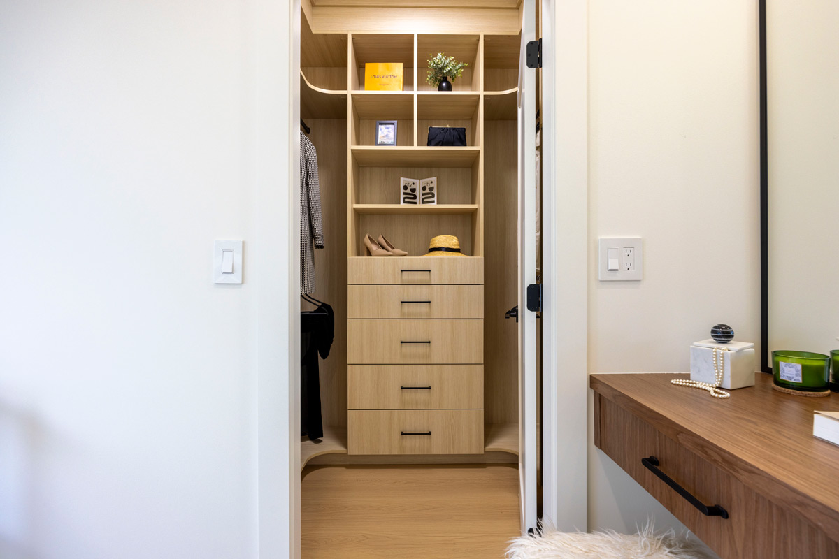 Built by Asthan Homes in Burnaby, BC, walk-in closet with light wood drawers, shelves, a hanging coat, and a vanity table beside the doorway.
