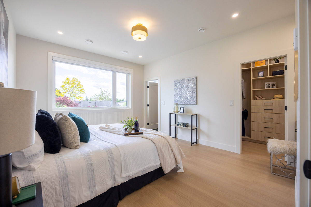 Built by Asthan Homes in Burnaby, BC, bright bedroom with large window, neatly made bed with pillows and tray, light wood floor, and open walk-in closet.