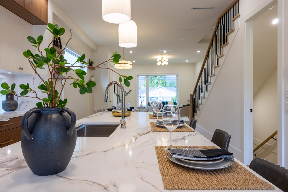 Built by Asthan Homes in Burnaby, BC, kitchen with marble island, black vase with green plant, table settings, and staircase in the background.