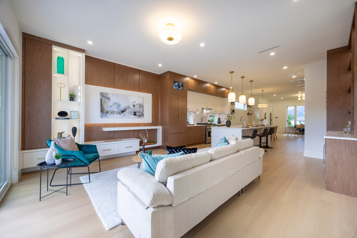 Built by Asthan Homes in Burnaby, BC, open living room and kitchen with white sofa, teal armchair, wooden cabinetry, and a kitchen island with pendant lights.