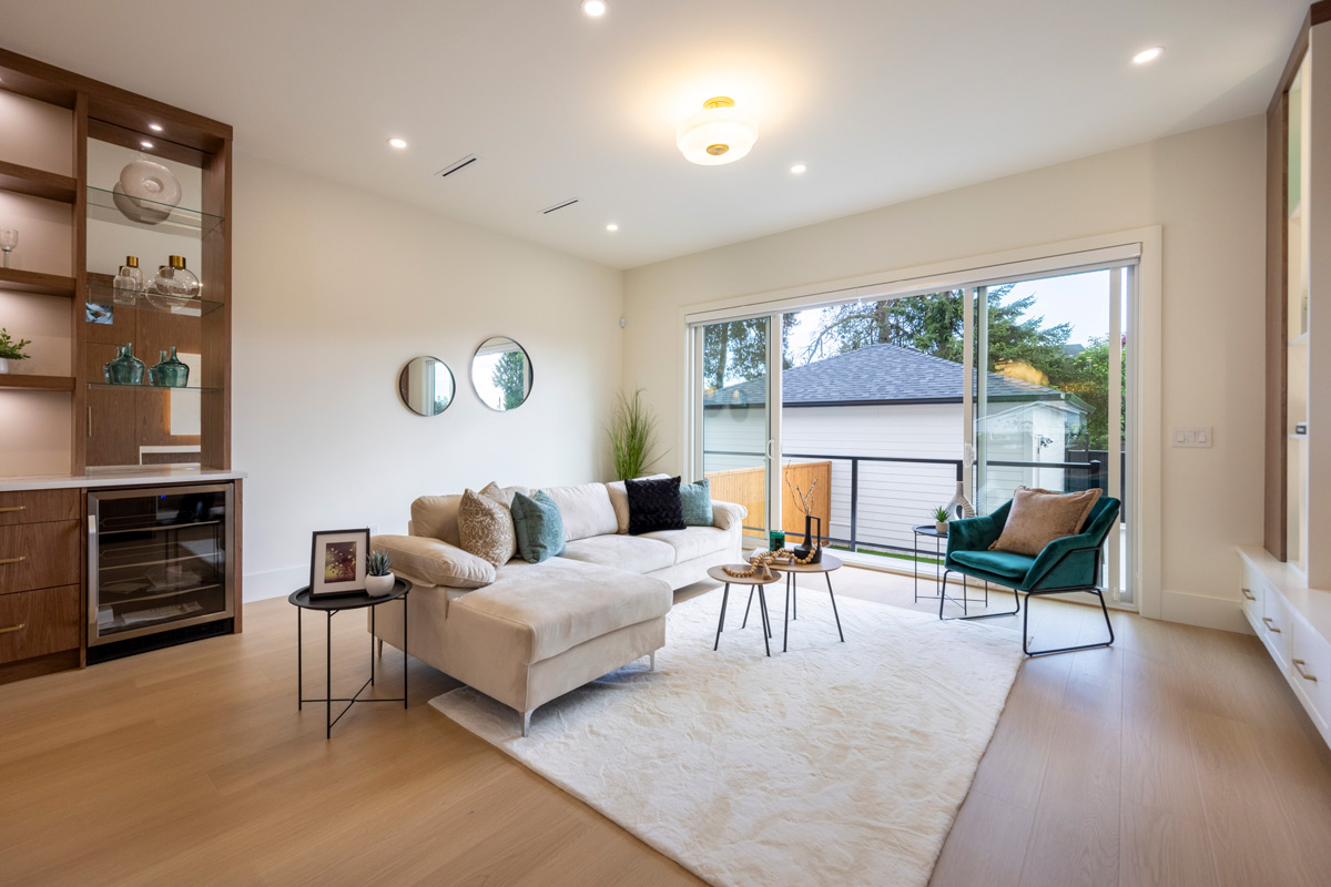 Built by Asthan Homes in Burnaby, BC, living room with beige sectional sofa, white shag rug, teal armchair, round coffee tables, and large sliding glass doors leading to an outdoor area.
