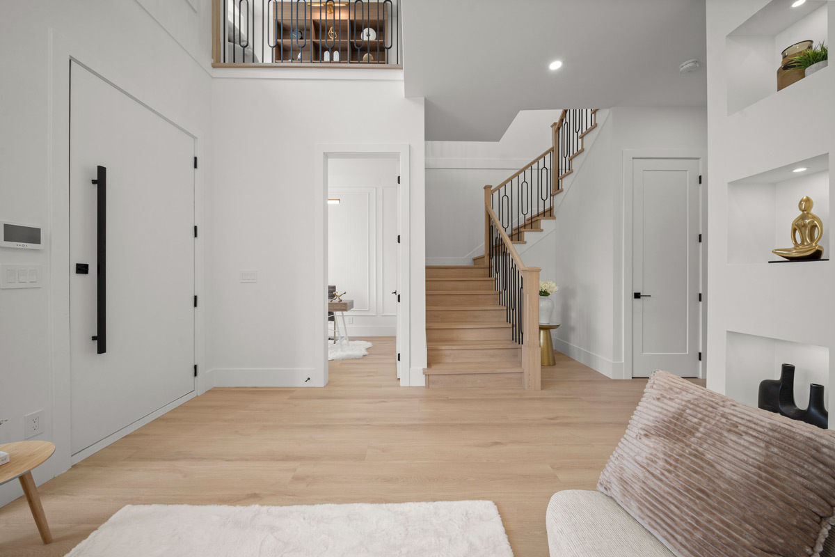 Built by Asthan Homes in Burnaby, BC, living room with light wood floor, staircase with wooden handrail and black balusters, white walls, and minimalistic decor including a fuzzy rug and plush pillow.