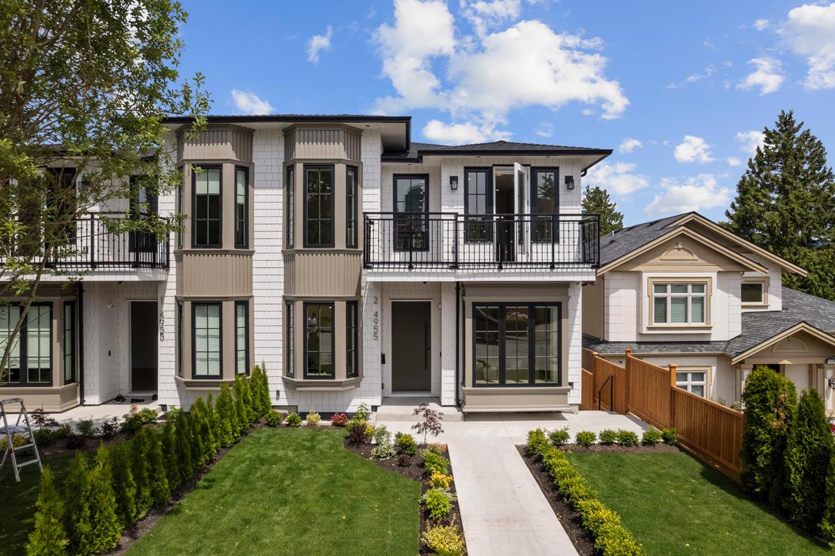 Built by Asthan Homes in Burnaby, BC, two-story duplex with white siding, bay windows, black window frames, and landscaped front yard with green grass and shrubs.