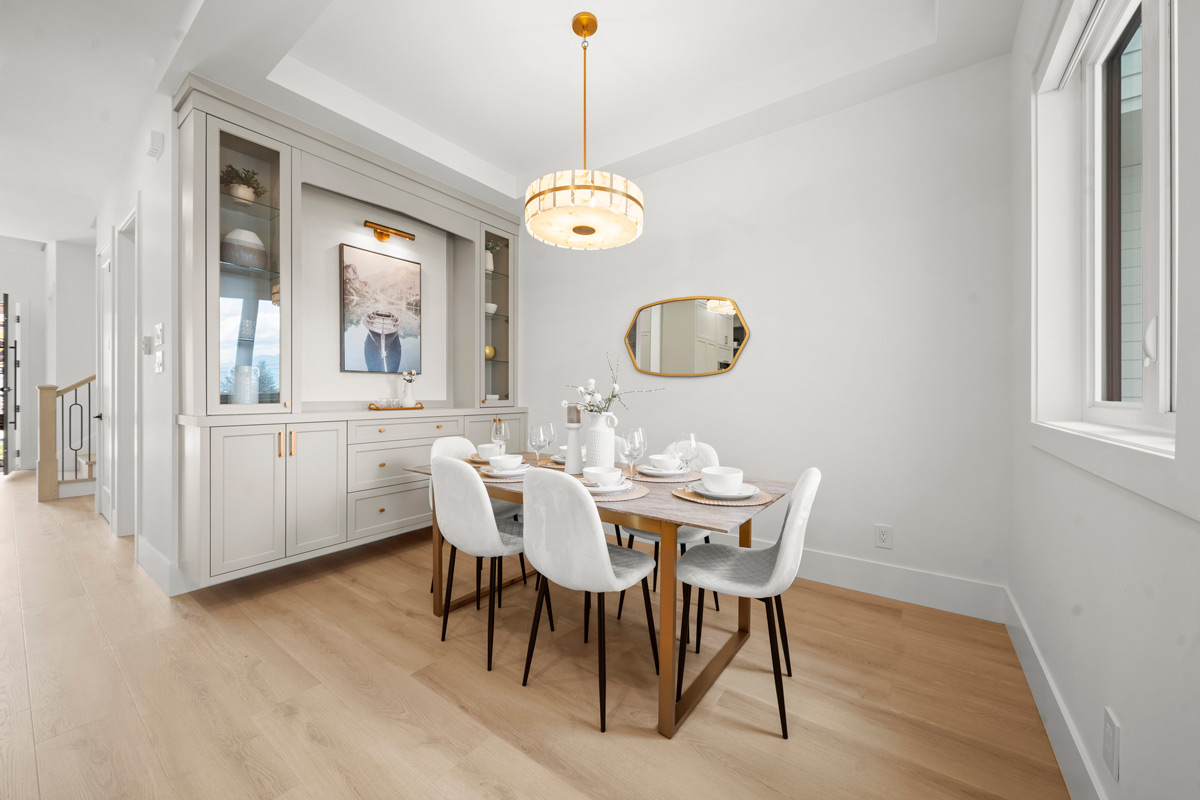 Built by Asthan Homes in Burnaby, BC, dining room with wooden floor, white chairs around a wooden table set with white dishes, a built-in cabinet with glass doors and decorative items, a pendant light, and a wall mirror.