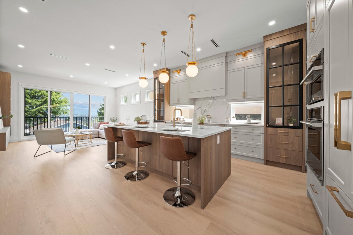 Built by Asthan Homes in Burnaby, BC, open kitchen with a large island featuring three brown bar stools, white marble countertop, light wood flooring, and pendant lights, opening to a living area with large windows overlooking trees and water.