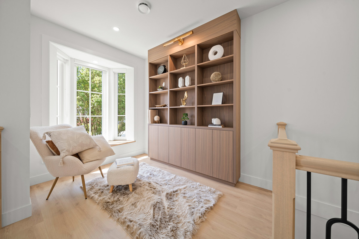 Built by Asthan Homes in Burnaby, BC, bright reading nook with a white armchair, matching ottoman on a fluffy rug, and a large wooden bookshelf with decorative items near a bay window.