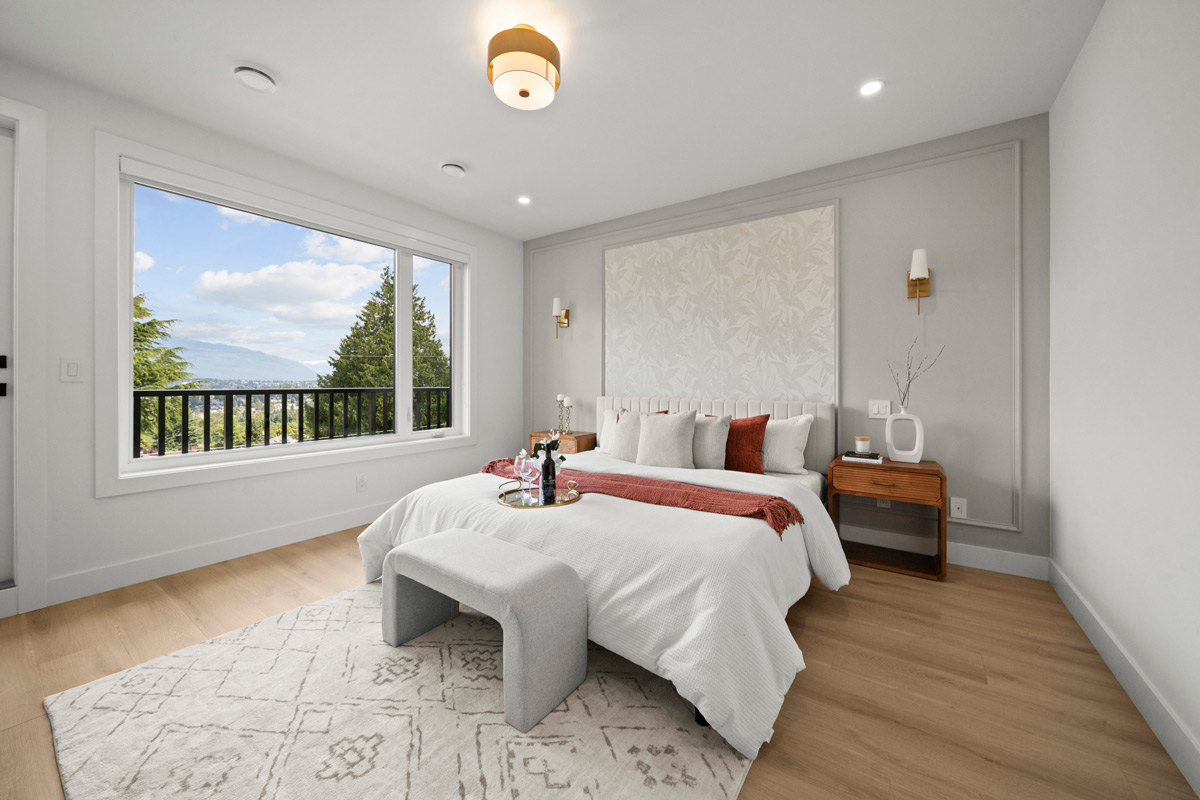 Built by Asthan Homes in Burnaby, BC, bedroom with a large window showing trees and mountains, bed with white bedding and red accents, wooden nightstands, and a light gray bench on a patterned rug.